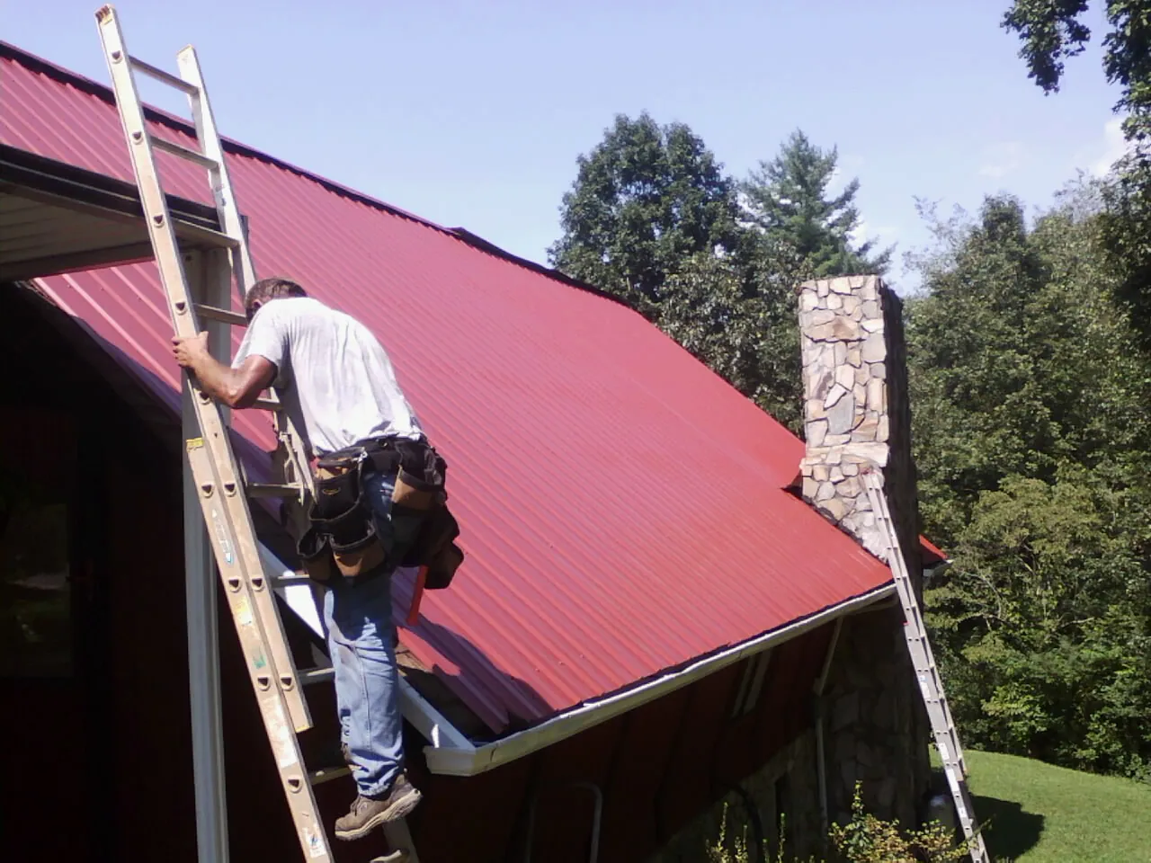 Roofer on a ladder beside a red metal roof and stone chimney — Soffit & Fascia Repair by Roofing Brothers in West Newbury